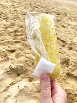 Boiled corn on the beach held in a hand, wrapped in plastic with salt packet Stock Photos