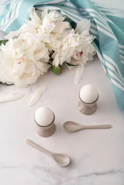 Boiled eggs in ceramic egg cups on background of beautiful white peony and tu Stock Photos