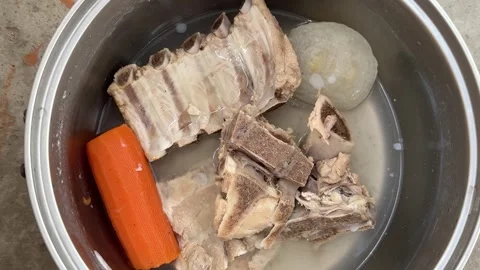 Boiled pork bones with a carrot and onion sit in water inside a steel pot Vídeos de archivo 341951874