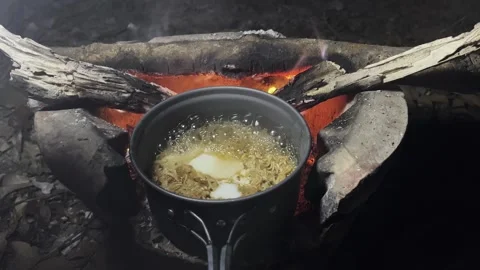 Boiling instant noodles and egg in a field pot on stove over a fire. Stock Footage 247399669