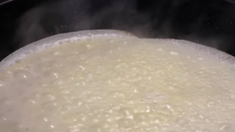 Boiling milk in a black saucepan with bubbles forming and steam rising Stock Footage 332423522