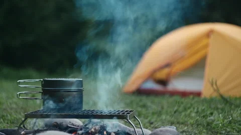 Boiling water over campfire on background tent and green forest in evening Stock Footage 247316544
