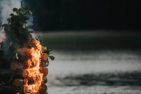 Bonfire with burning logs and green foliage near a calm lake, smoke rising .. Stock Photos