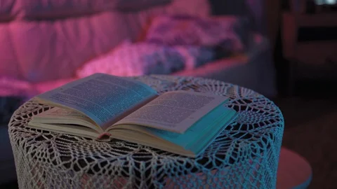 Book is lying on the table in a colored light.behind her is TV that doesn't work Stock Footage 128458386