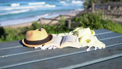 Book, shawl and a hat on the table with blue ocean waves in background. Stock Footage 134505135