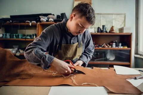 In a boot shop, an experienced young master cuts out leather shoe elements with Stock Photos