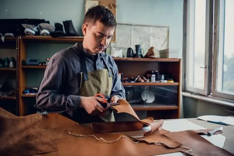In a boot shop, an experienced young master cuts out leather shoe elements with Stock Photos