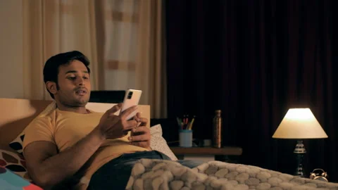Bored Indian guy in a yellow T-shirt and blue jeans using mobile before sleeping Stock Footage 155261948