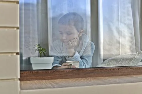 A bored kid at the window thoughtfully sits with a mug. Stock Photos