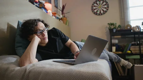 Bored Man with Glasses Lying in Bed Using Laptop for Homework Stock Footage 338423455