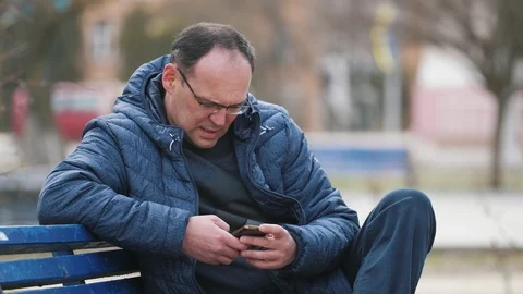 Bored man in glasses siting on a bench and  using smartphone on the street in Stockbeeldmateriaal 105343193
