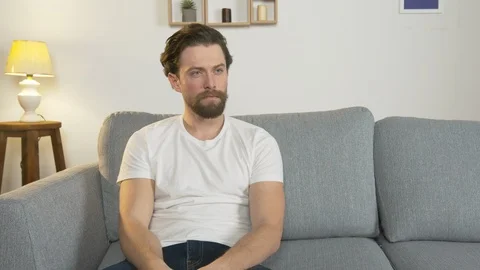 Bored man sits alone on a sofa in a room and watches TV, holds a remote control Video stock 127412590