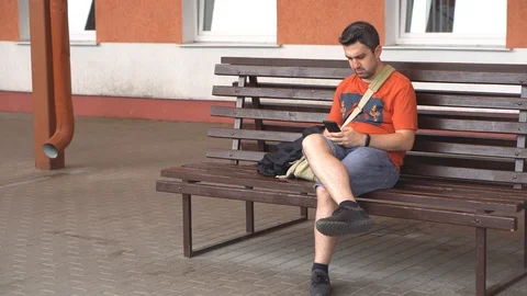 Bored man sitting on bench outside waiting for and calling on a phone that has Stock Footage 111861699