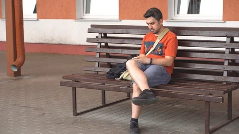 Bored man sitting on a bench outside waiting for photo on his phone Stock Footage 111862513