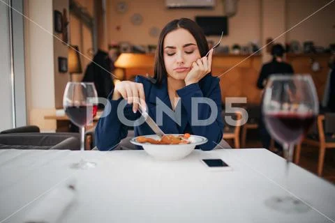 Bored young woman sit at table in restaurant alone. She look at food and touch Stock Photos