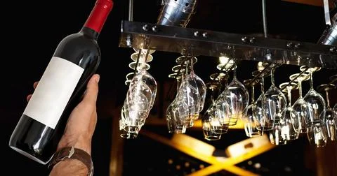 Bottle of red wine in hand on background of wine glasses hanging in the bar o Foto stock