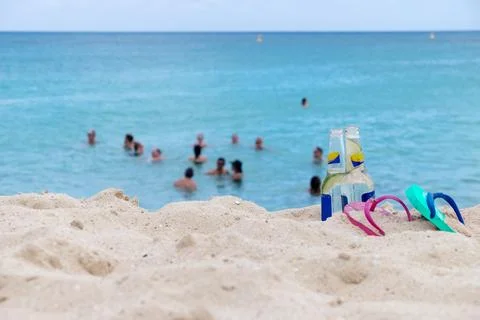 Bottles with lime and flip-flops on beach, people swimming in clear ocean Stock Photos