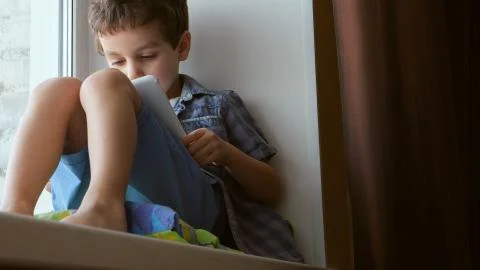 BOTTOM VIEW: Cute little boy uses a white tablet PC on a windowsill at home Stock Photos