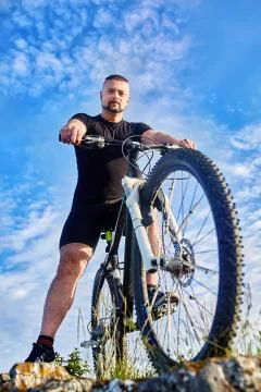 Bottom view of young cyclist standing with bike on rocks against blue sky. Stock Photos