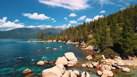 Boulder beach and pine covered slopes beside Lake Tahoe. People gather near pale Stock Footage 346724883