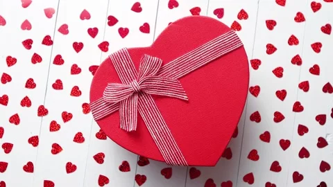 Boxed gift placed on heart shaped red sequins on white wooden table Stock Footage 228605513