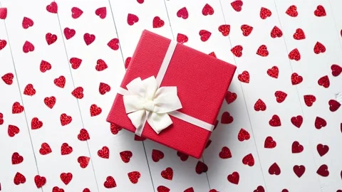 Boxed gift placed on heart shaped red sequins on white wooden table Stock Footage 228863320