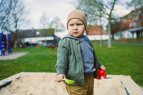 A boy of 2 years old in a spring jacket and hat plays with sand in a sandbox. Stock Photos