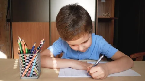 A boy of 5-6 years old at the table writes letters with a pen in a notebook in Stock Footage 290495358