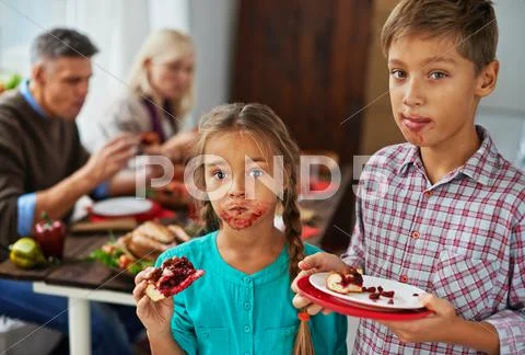 Boy and girl eating sweet pie with dirty mouths, their parents eating in the bac Stock Photos
