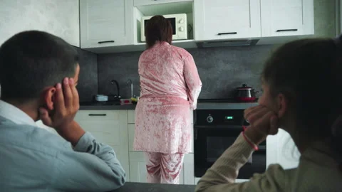 Boy and girl sit at kitchen table watching woman in pink robe prepare food in Stock Footage 317929079