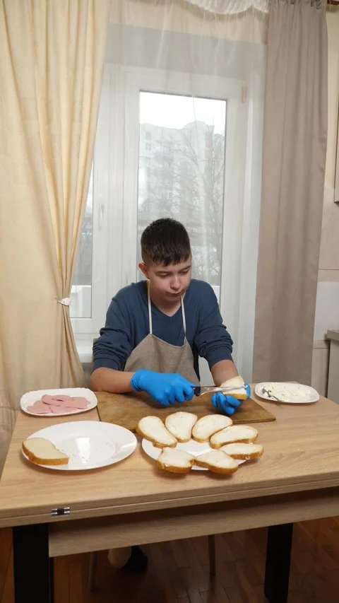 Boy in apron and blue sweater spreads cream cheese on bread with spoon while  Stock Footage 337225623