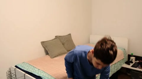 Boy arranges pillows on a made bed, leaning forward in a wide side view with Stock Footage 327927177