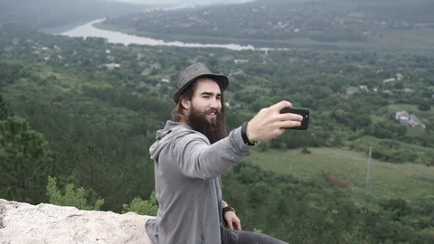 A boy with a beard and hat takes a selfie at a beautiful place near a river. Stock Footage 127184068