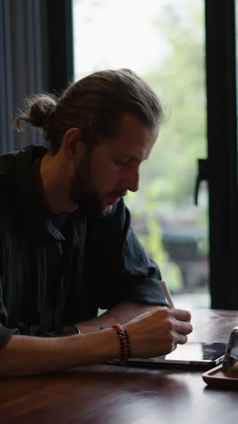 Boy with beard and ponytail draws something on his tablet Stock Footage 265475622