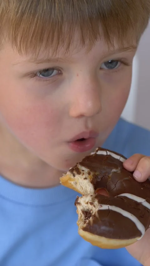 Boy biting chocolate donut with focused expression, food concept for parenting Video stock 333680380