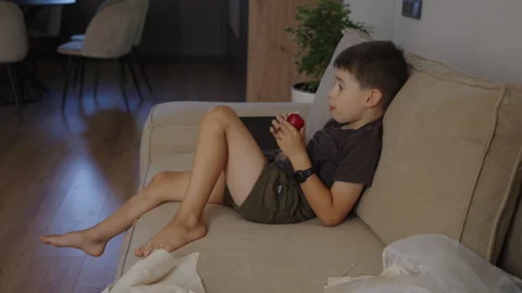 Boy bitting red apple while watching cartoon on TV, relaxing at home after back Stock Footage 249791837