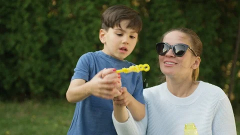 Boy blowing bubbles while mother helping him by holding the wand in backyard Stock Footage 119989733