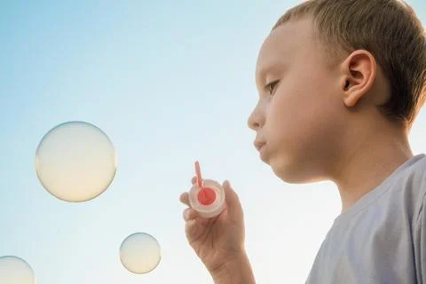 Boy blowing soap bubbles on the evening blue sky background. Side view. Stock Photos