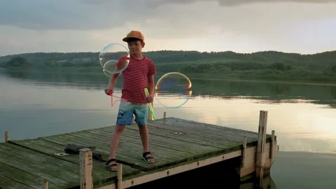 A boy blows huge bubbles at sunset on the pier over the lake Stock Footage 206634541