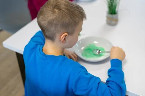 A boy in a blue jacket making green slime at a table in a white plate Stock Photos