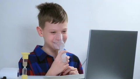 Boy is breathing in mask using nebulizer for inhalation at home watching laptop. Video stock 152714717