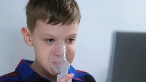 Boy is breathing in mask using nebulizer for inhalation at home watching laptop. Video stock 159484716