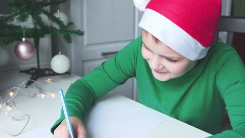A boy in bright clothes and a cap sitting at the table writes a Christmas letter Stock Footage 165384023