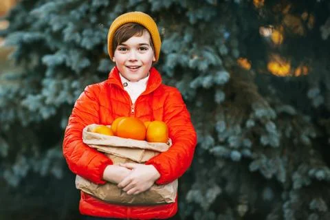 A boy in a bright orange jacket and a yellow hat holds a large package with Stock Photos