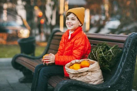 A boy in a bright orange jacket and a fashionable yellow hat sits on a bench Stock Photos