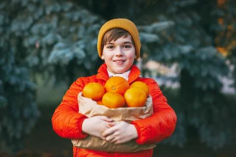 A boy in a bright orange jacket and a yellow hat holds a large package with Stock Photos