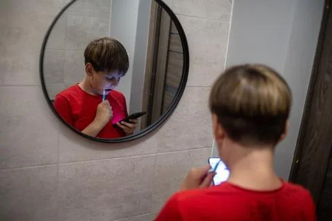 Boy Brushing Teeth While Using Smartphone In Mirror Stock Photos