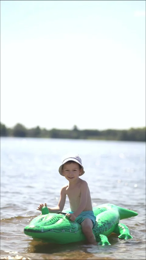 Boy in a bucket hat sits smiling on a green inflatable crocodile float Stock Footage 341989151