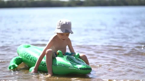 Boy in a bucket hat sits smiling on a green inflatable crocodile float Stock Footage 341989163