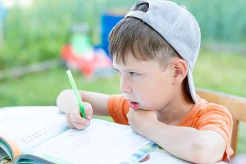 A boy in a cap solves math examples at a table on the street. Child doing hom Stock Photos